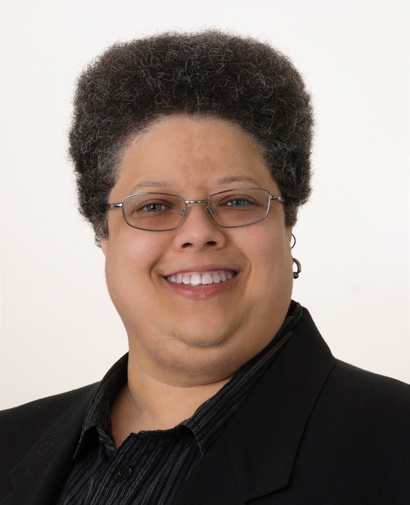 Professional headshot of a smiling woman wearing glasses and a black blazer against a light background.